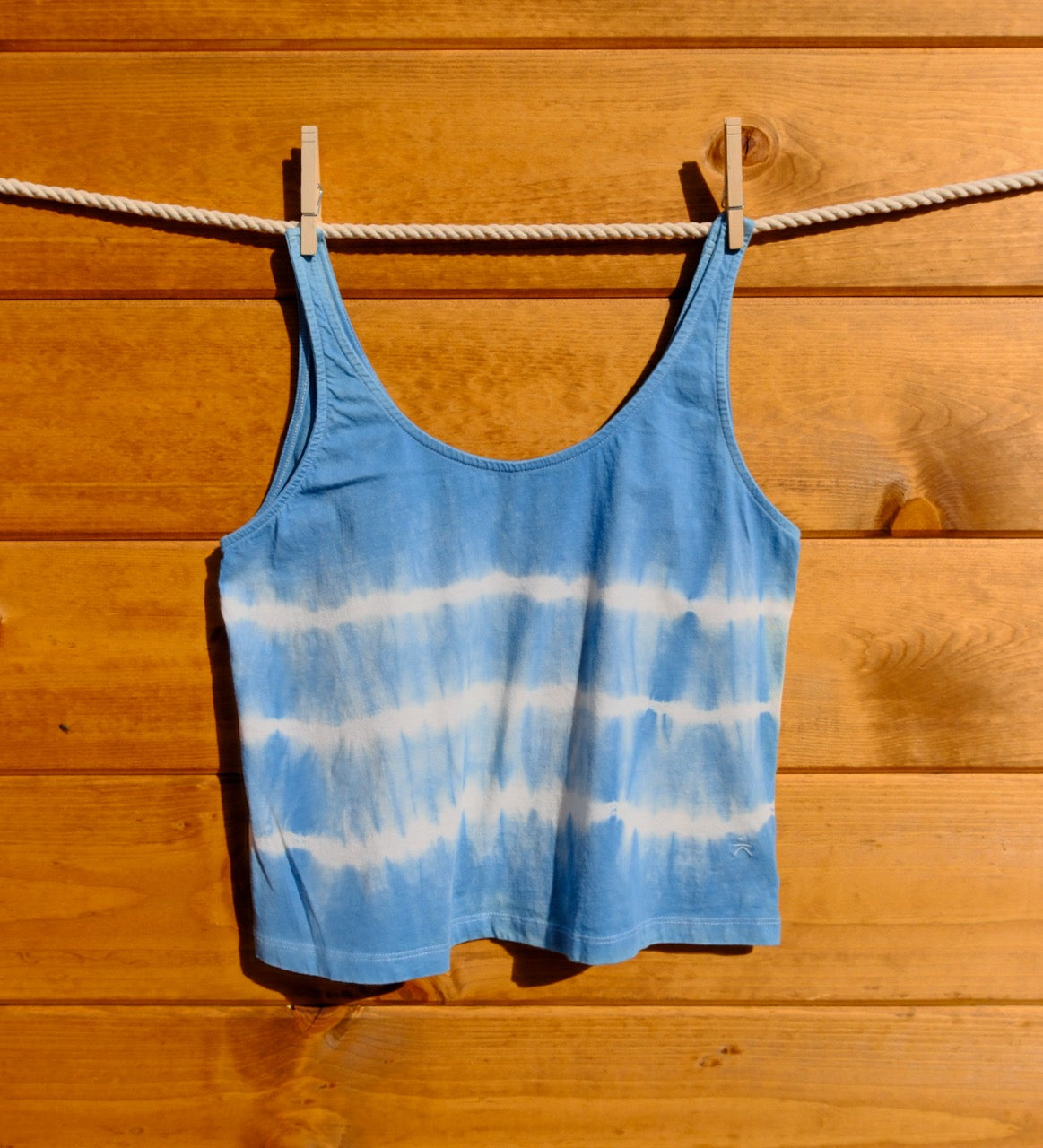 Blue tie-dye tank top hanging on a clothesline against a wooden wall.