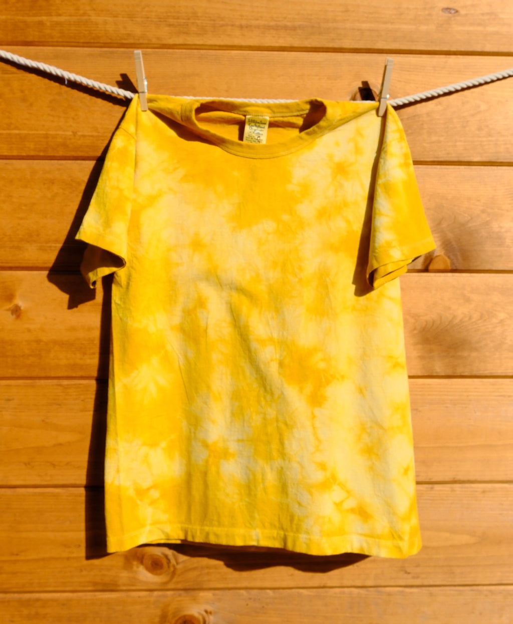 Yellowish orange tie-dye shirt hanging on a clothesline against a wooden wall.
