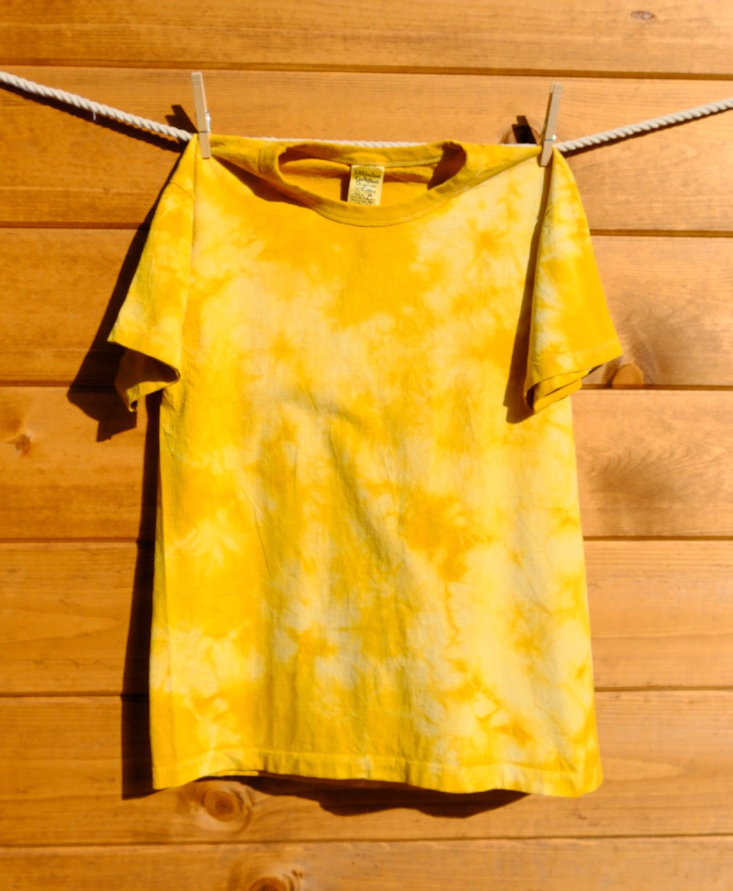 Yellowish orange tie-dye shirt hanging on a clothesline against a wooden wall.
