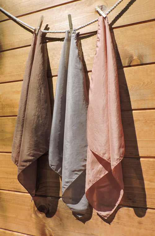 Three silk scarves in brown, gray, and pink hanging on a clothesline against a wooden wall.